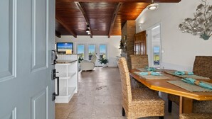 A spacious dining area with a high ceiling and exposed beams, a long wooden table with woven chairs, and a view of the outdoors through large windows. - Sunset Vista | Private Dock, Dog-Friendly Home | My Beach Getaways (Pensacola Beach)