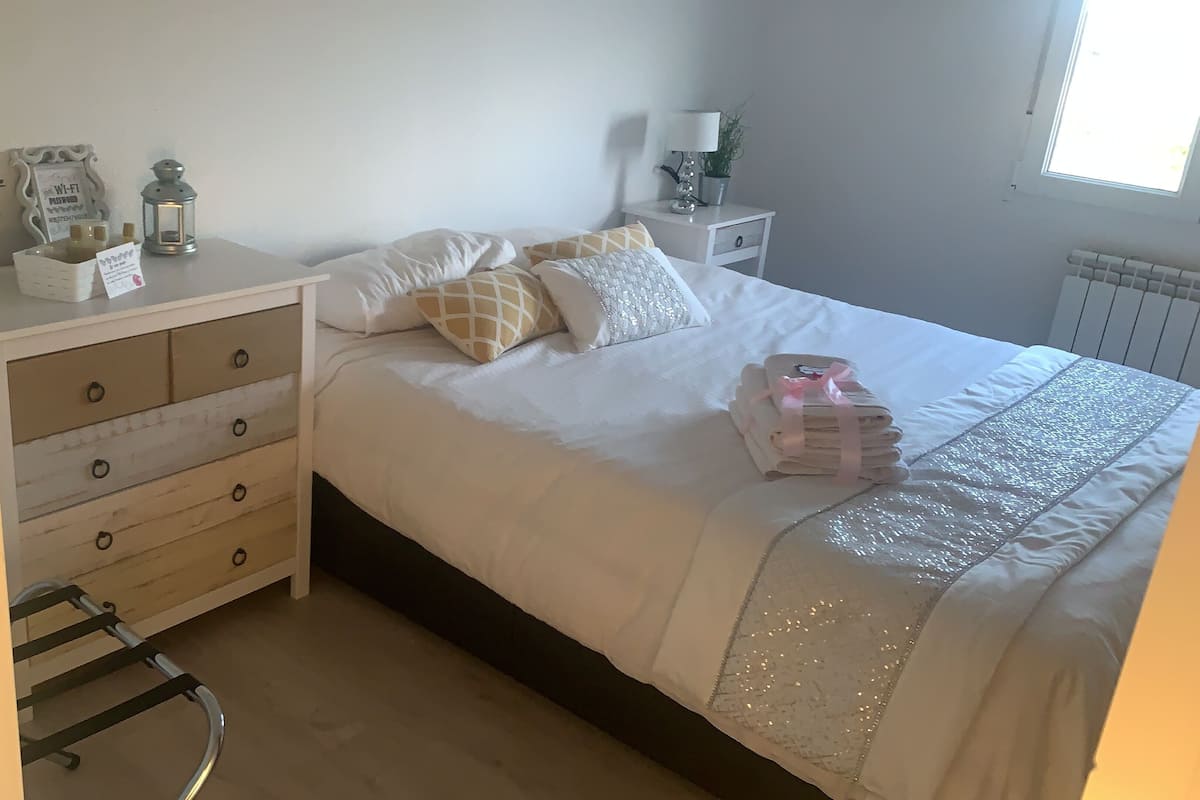 A neatly made bed with a white comforter and decorative pillows, a bedside table with a lamp and a small plant, and a wooden dresser with drawers.