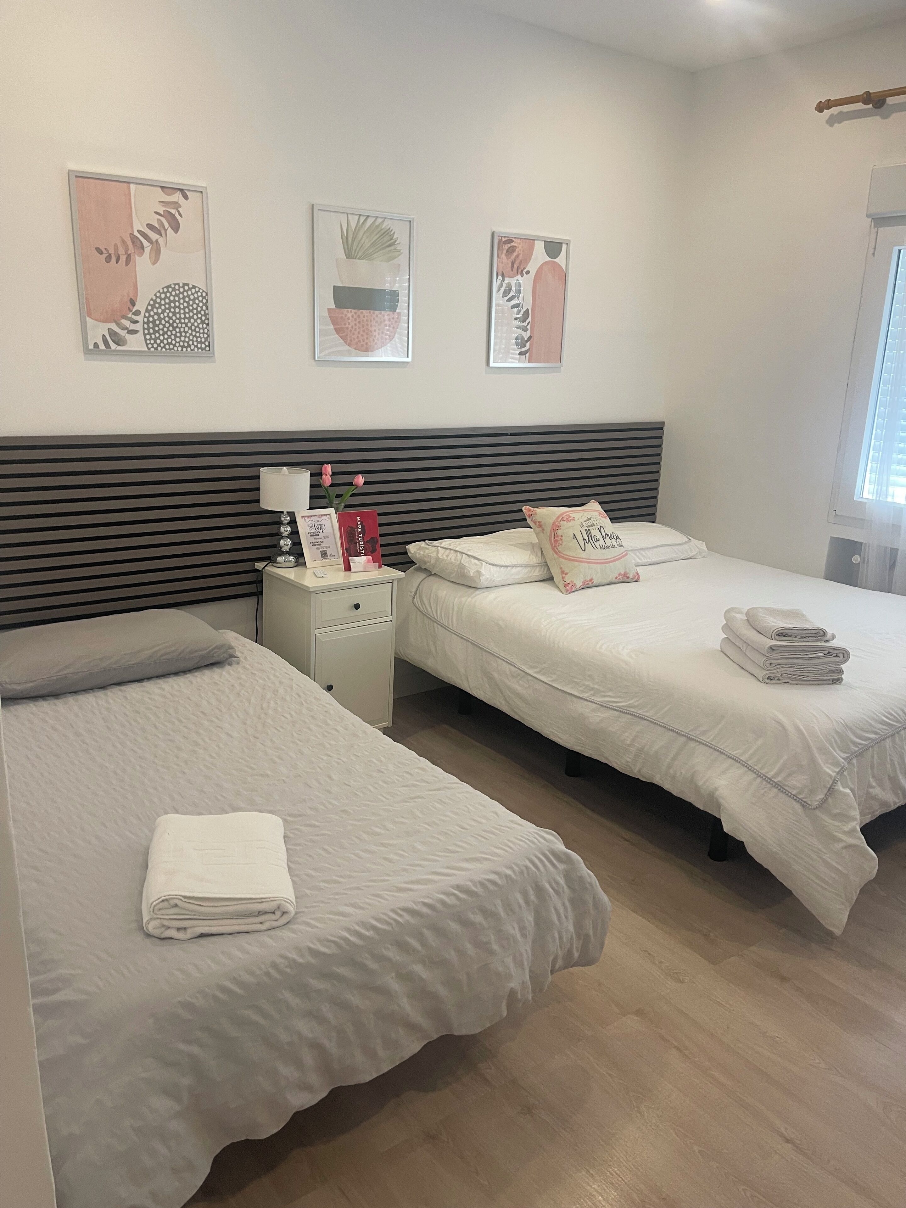 A hotel room with two single beds, a nightstand with a lamp, a small table with a vase and a book, and three wall art pieces.