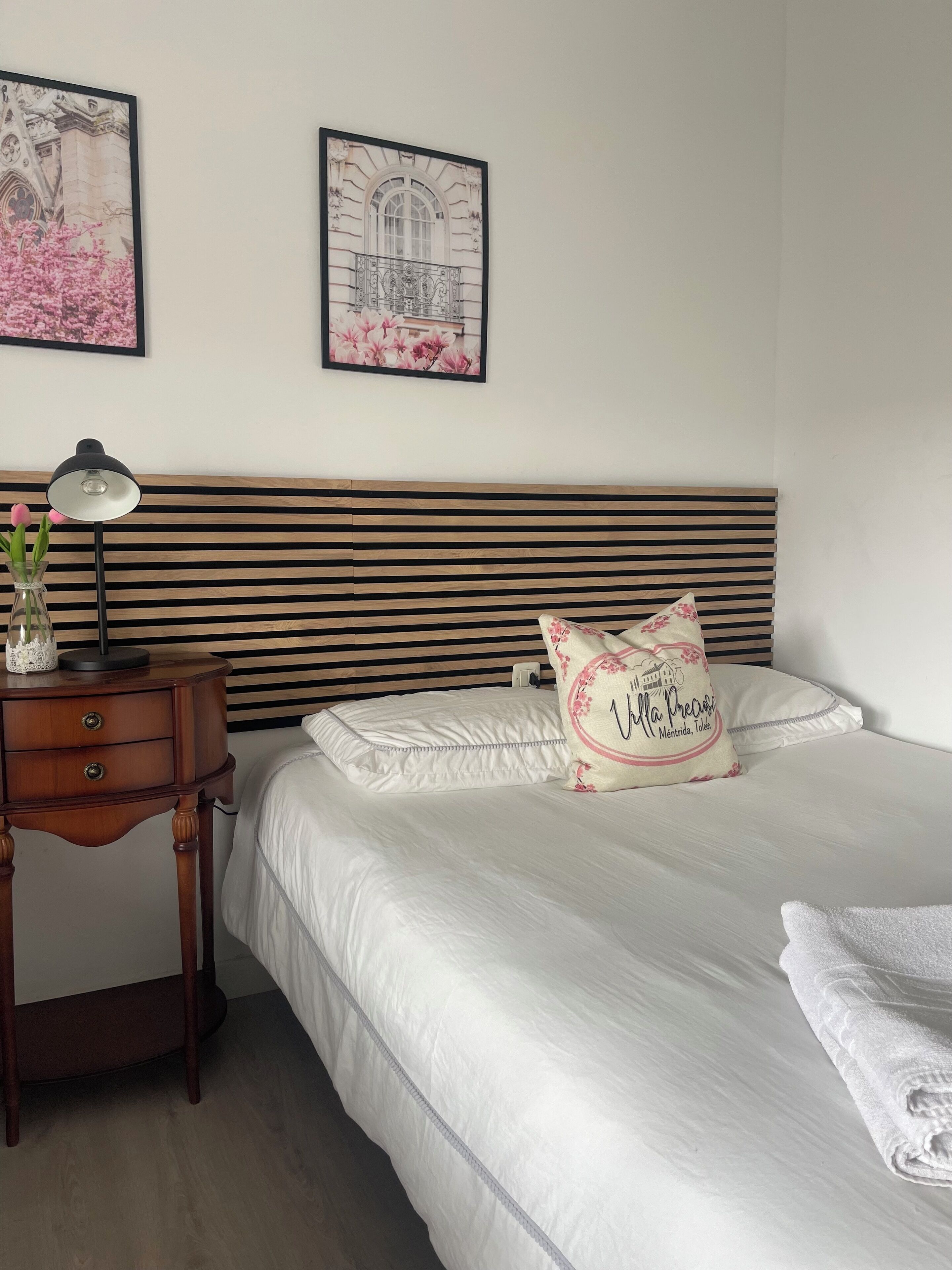 Una cama bien hecha con sábanas blancas, una lámpara de noche, un florero con flores y un cojín decorativo con texto.