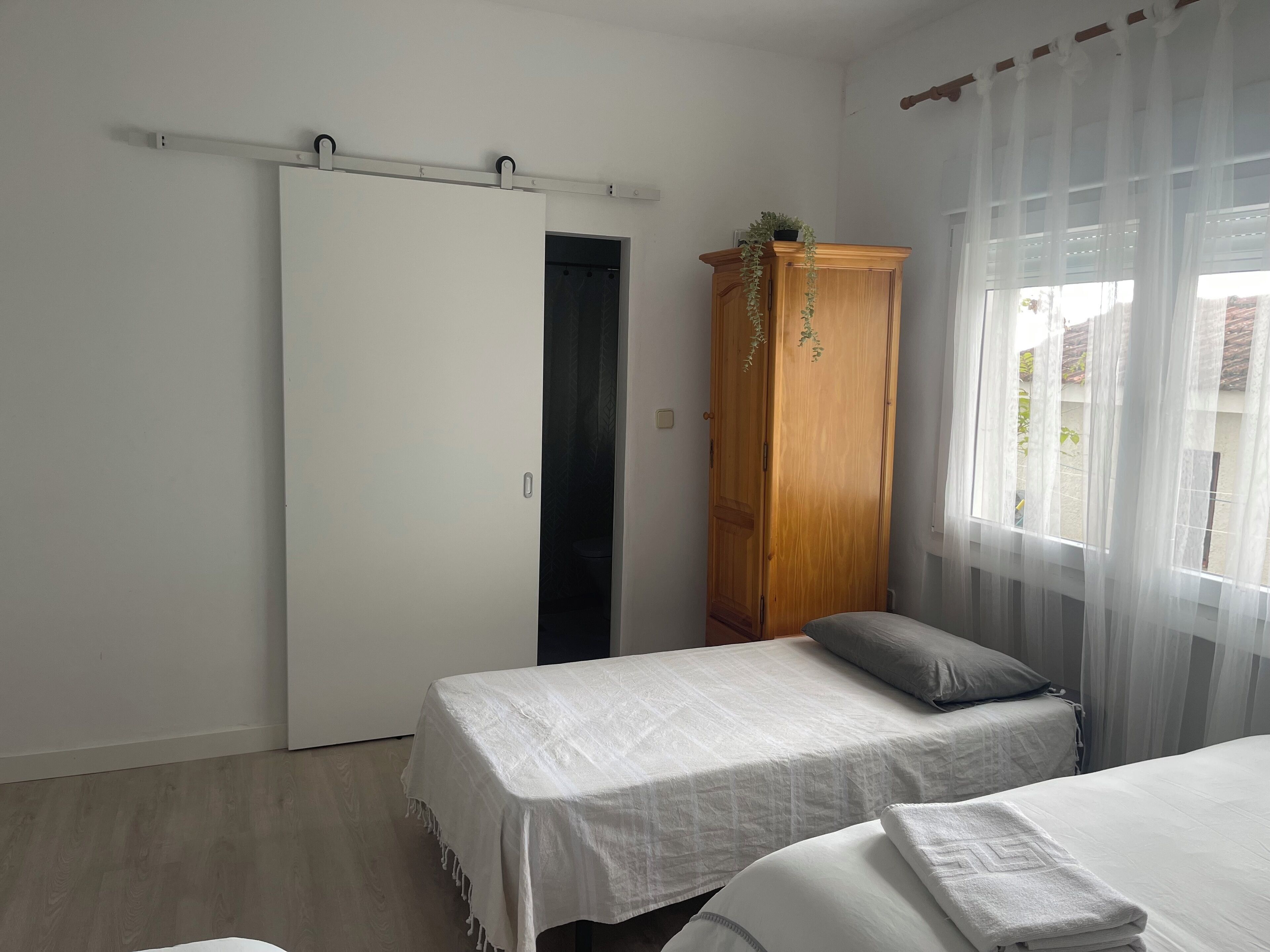 Un dormitorio con dos camas, un armario y una ventana con cortinas translúcidas.
