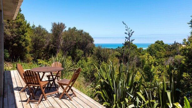 A wooden deck with a round table and chairs overlooking a coastal landscape. - Secluded Haven with Stunning Hokianga Harbour Views (Omapere)