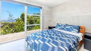 A modern bedroom with a large bed, a nightstand, and a view of greenery through a sliding glass door. - Secluded Haven with Stunning Hokianga Harbour Views (Omapere)