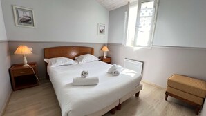 Ein ordentlich bezogenes Bett mit weißen Leinen, zwei Nachttischlampen, ein kleiner Nachttisch aus Holz, ein Fenster mit Jalousien und ein Korbstuhl.