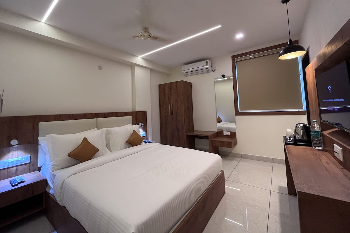 Quarto de hotel moderno com uma cama bem arrumada, uma mesa de cabeceira de madeira, uma escrivaninha com cadeira, um espelho grande e um ventilador de teto.