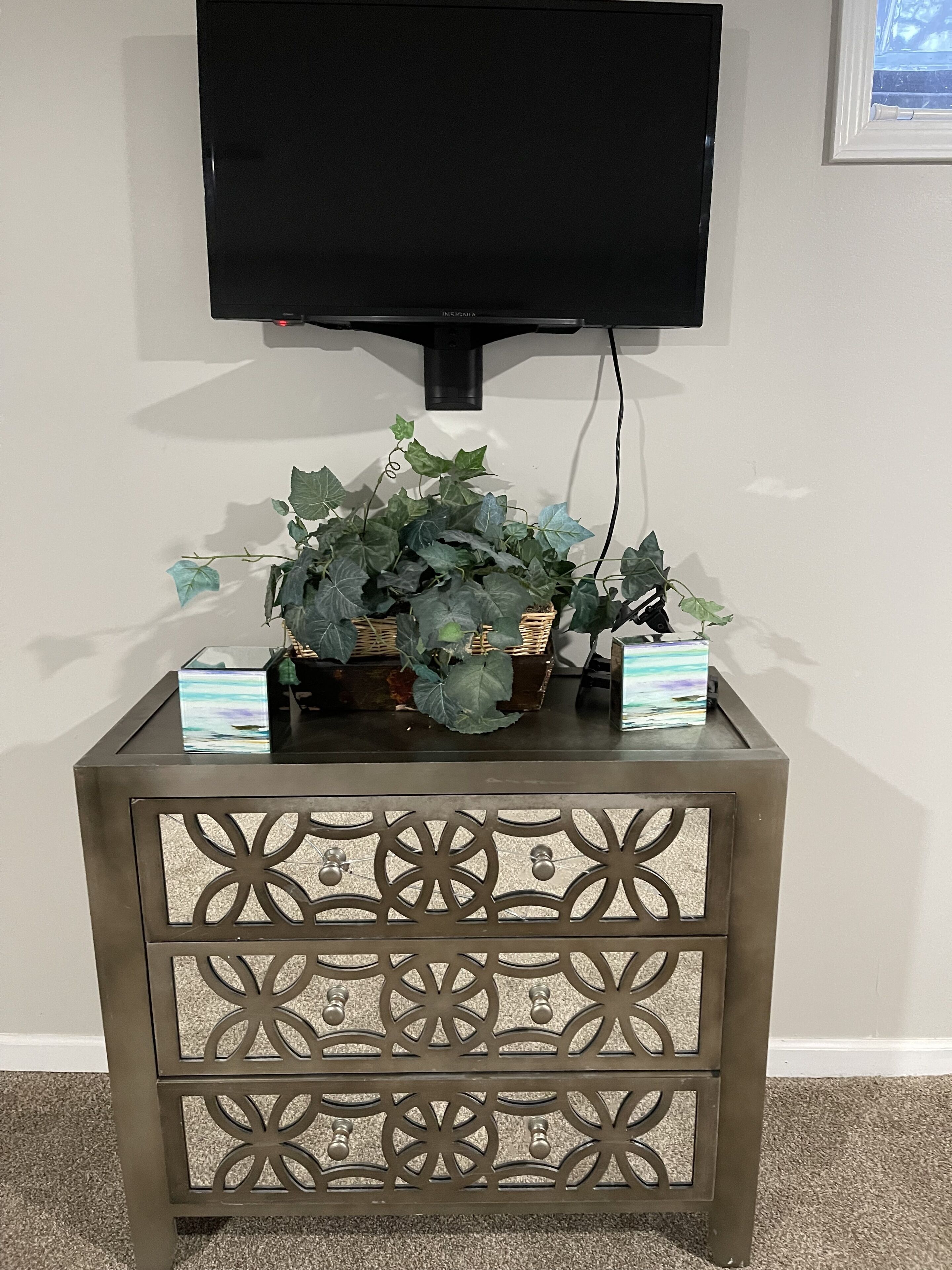 Un téléviseur fixé au mur, au-dessus d’une petite table d’appoint décorative, sur laquelle se trouvent un panier en osier et une plante.