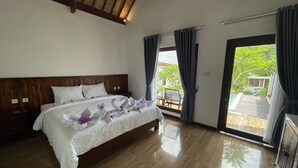 A hotel room with a wooden headboard, a large bed with white linens, a wooden floor, and a view of a pool and greenery through the window. - THELOCAL AREGULING  (Kuta)