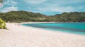 A beach with clear turquoise water, sandy shore, and lush green hills in the background. - THELOCAL AREGULING  (Kuta)