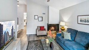 Una sala moderna con chimenea, un sofá azul, una mesa de centro con un florero y una televisión que muestra un paisaje urbano.