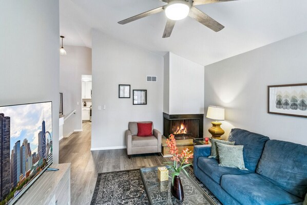 A modern living room with a fireplace, a blue sofa, a coffee table with a vase, and a cityscape TV.
