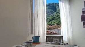 Ein runder Glastisch mit weißer Tischdecke, eine blaue Karaffe und eine weiße Tasse auf einem Balkon mit weißen Vorhängen und Blick auf einen bewaldeten Hügel.