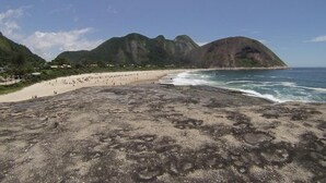 Ein Strand mit felsigem Vordergrund, Sandstrand und Bergen im Hintergrund.
