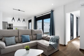 Un salon moderne avec un canapé d’angle, des luminaires suspendus, une cuisine en arrière-plan et de grandes fenêtres avec des rideaux.