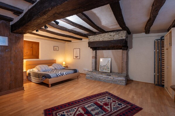 A bedroom with a bed, wooden beams, a stone fireplace, and a red patterned rug.
