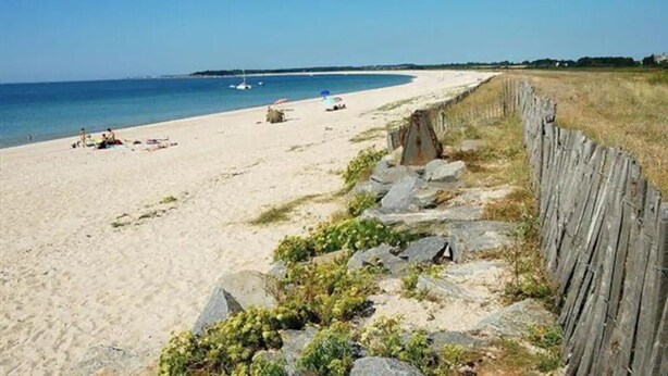 Une plage de sable bordée d’une clôture en bois, un voilier sur l’eau et un ciel dégagé.