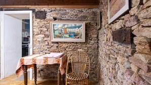 Une pièce rustique avec un mur en pierre, un tableau encadré, une table recouverte d’une nappe à motifs floraux et une chaise en osier.