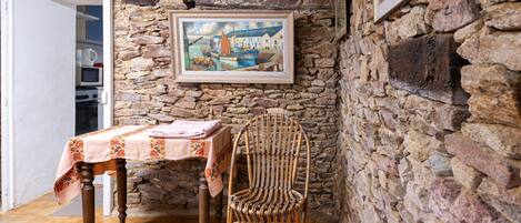 Una stanza rustica con parete in pietra, un quadro incorniciato, un tavolo con tovaglia floreale e una sedia in vimini.