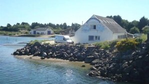 Un bâtiment situé sur la côte, avec un rivage rocheux, un petit quai et un fourgon blanc garé à proximité.