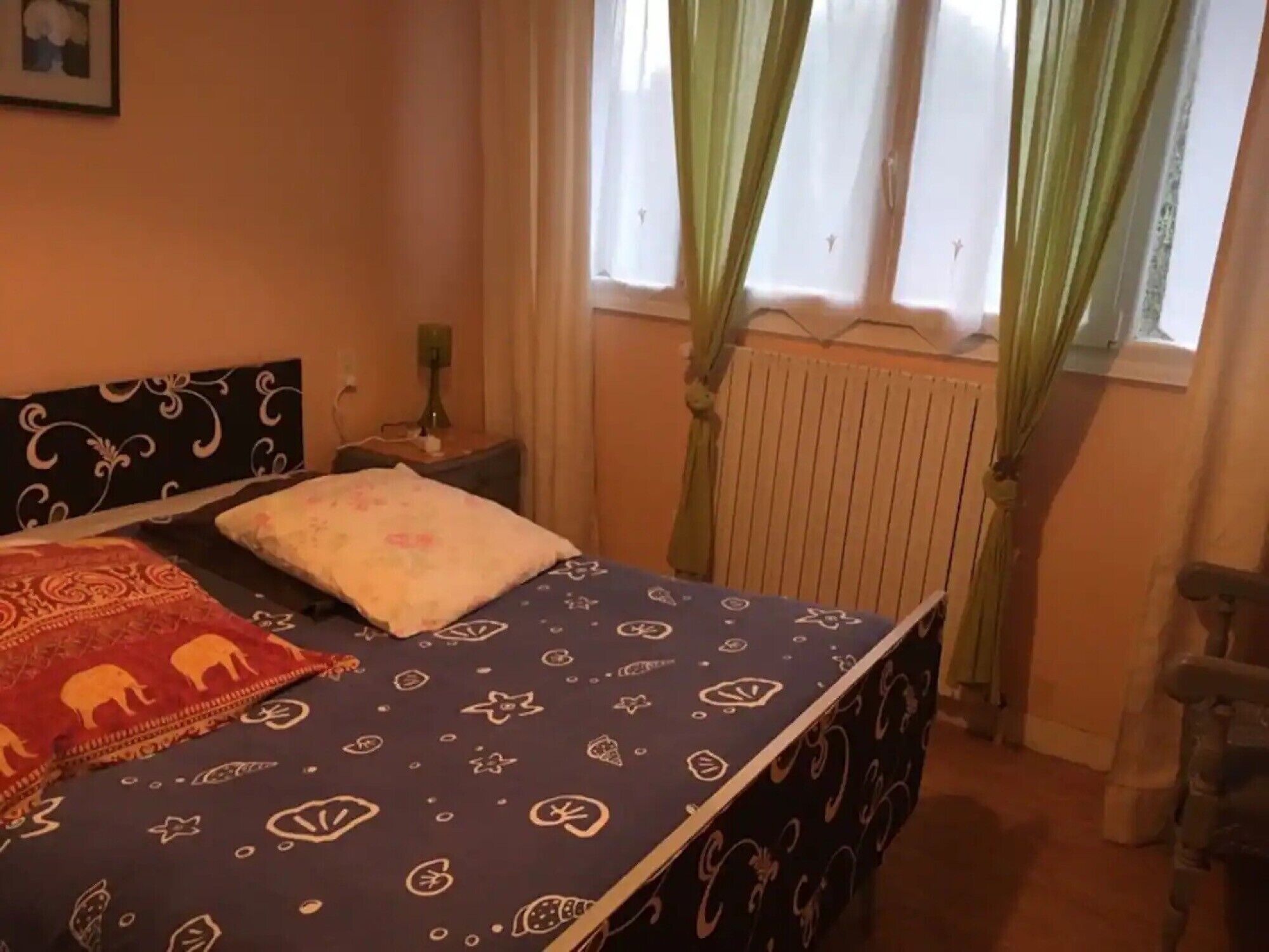 Un dormitorio con cama, mesita de noche, radiador y ventana con cortinas.