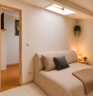 Une chambre à coucher avec un lit, une table de chevet avec une lampe, une plante suspendue et une porte donnant sur une autre pièce.