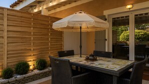 En udendørs terrasse med et bord, stole, parasol, lyskæder og et træhegn.