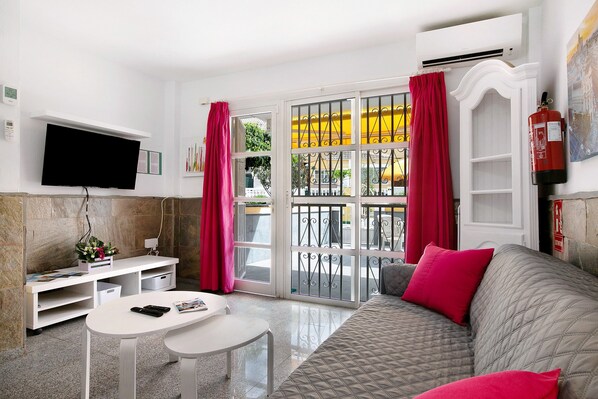 Un salon comprenant un canapé, un téléviseur, une petite table, une console blanche et une fenêtre avec des rideaux.