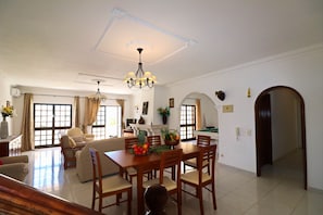 A spacious dining area with a wooden table, chairs, and a chandelier. There is a sofa and a window with curtains in the background.