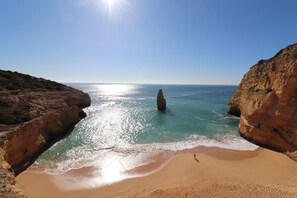 A beach with a clear sky, a rocky cliff, and a person standing on the sand.