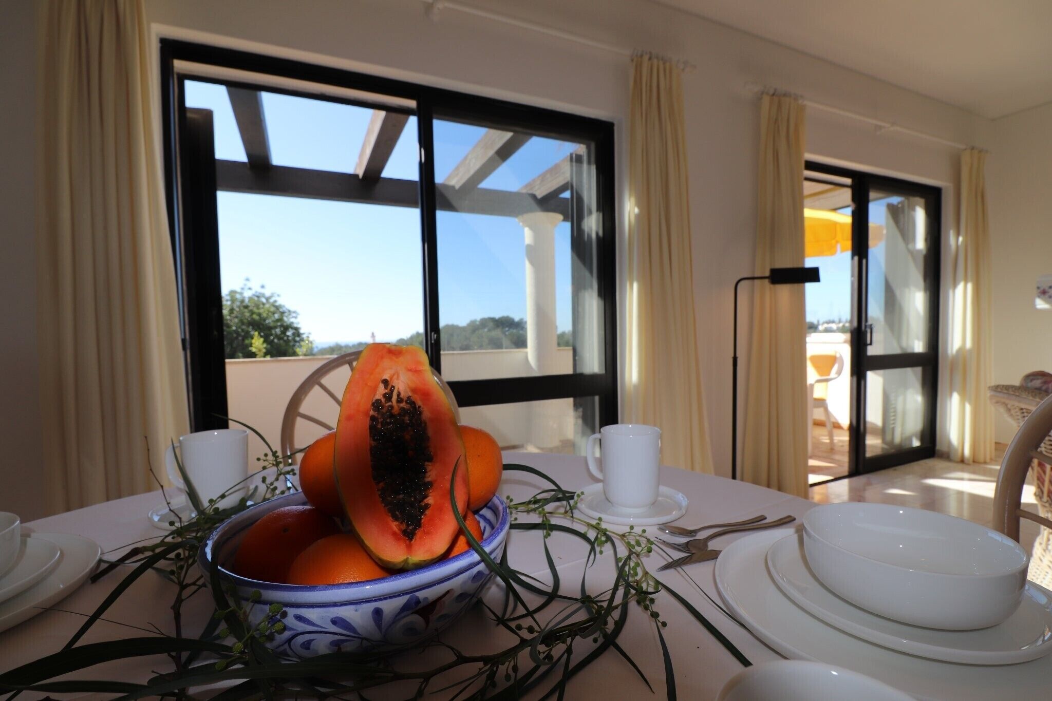 A dining table with a bowl of fruit, white plates, and cups, set against a backdrop of a window with a view of the outdoors.