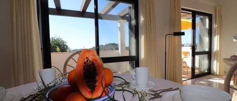 A dining table with a bowl of fruit, white plates, and cups, set against a backdrop of a window with a view of the outdoors.