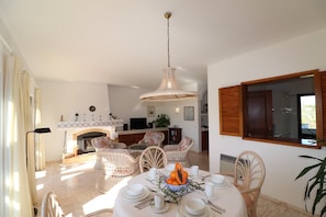 Un coin repas avec une table ronde dressée pour quatre personnes, entourée de chaises en osier, une cheminée et une cuisine en arrière-plan.