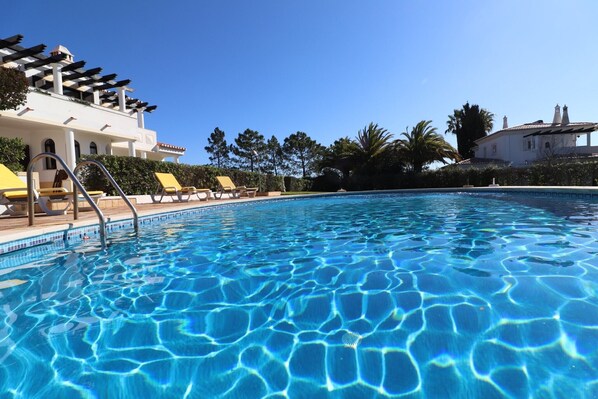 A swimming pool with clear blue water, surrounded by lounge chairs and a pergola.