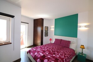 Un dormitorio con cama, ventana, puerta, luz empotrada en la pared y mesita de noche con lámpara.