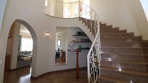 Eine geschwungene Treppe mit dekorativer Geländer, ein Holzboden und Blick in einen Wohnbereich mit Sofa und Essbereich.