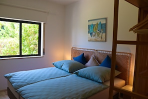 Ein Doppelbett mit einem gewobenen Kopfteil, ein Fenster mit Blick auf Grünflächen und ein Gemälde von Booten an der Wand.