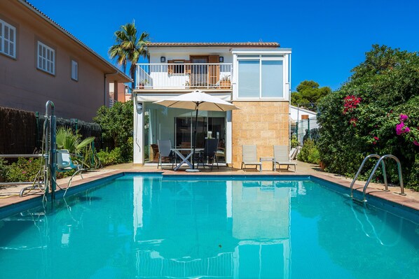 A swimming pool with a sun lounger, a covered seating area, and a two-story building with a balcony.