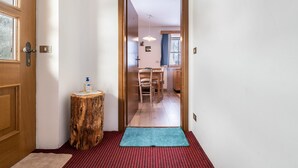 Un corridoio con una porta in legno, uno sgabello di legno con una bottiglia, un tappeto a righe rosse e nere, e una vista su una stanza con una sedia e un tavolo.
