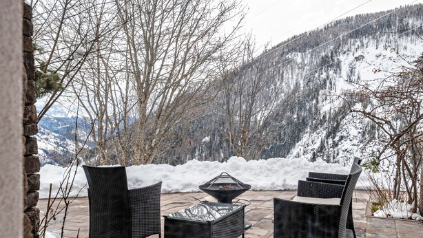 Un patio exterior con mobiliario de mimbre y una mesa con cubierta de vidrio, rodeado de montañas cubiertas de nieve.