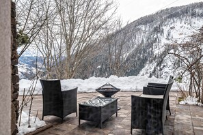 Un patio exterior con mobiliario de mimbre y una mesa con cubierta de vidrio, rodeado de montañas cubiertas de nieve.