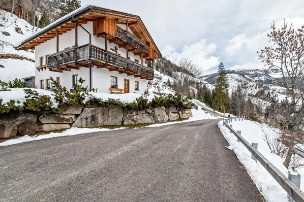 Un chalet de dos pisos con balcones, un muro de contención de piedra y una montaña nevada al fondo.