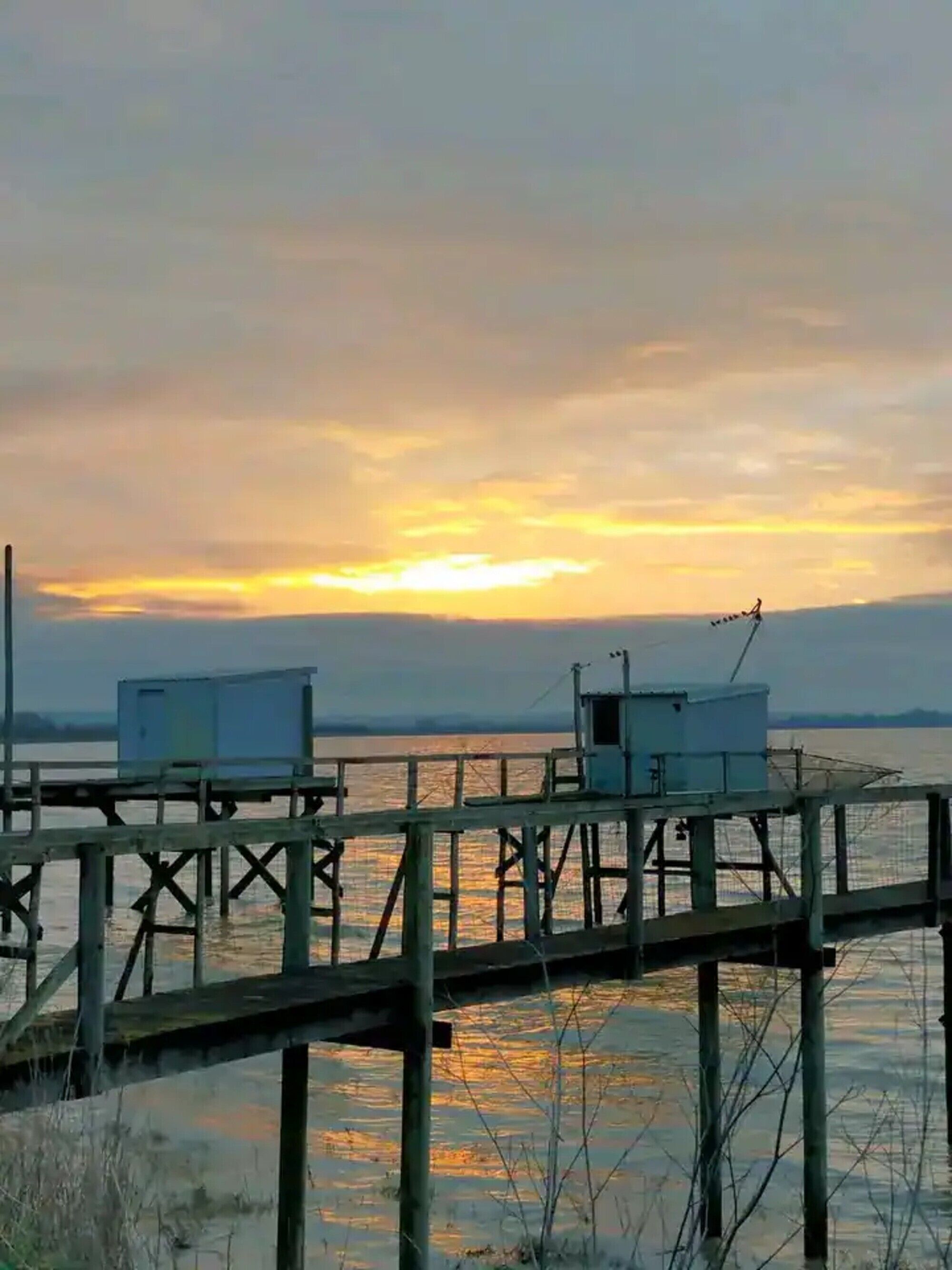 Un muelle de madera se adentra en aguas tranquilas, con dos pequeñas construcciones, probablemente cabañas o almacenes, y un cielo despejado.