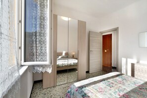 A bedroom with a bed, a wardrobe, a mirror, and a window with lace curtains.