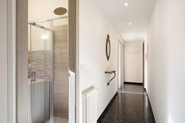 Un couloir avec une cabine de douche en verre, un radiateur et une étagère fixée au mur.