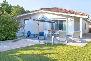Una casa con techo de tejas, un porche cubierto con sillas y una mesa, y un césped.