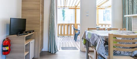 A room with a TV, a fire extinguisher, a wooden desk, a table with a tablecloth, and a view of the outside deck.