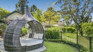 A curved, woven outdoor seating area with cushions and a table, surrounded by a well-maintained lawn and a black metal fence. - Experience Lavender Cottage, charming, cozy hideaway set in pretty bushland.  (Leura)