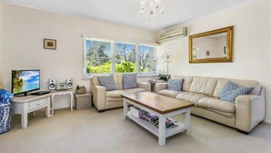 A living room with a beige sofa, a wooden coffee table, a TV, speakers, and a chandelier. - Experience Lavender Cottage, charming, cozy hideaway set in pretty bushland.  (Leura)