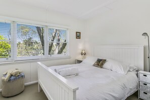 Ein Schlafzimmer mit einem weißen Bett, einem Nachttisch, einer Lampe, einem Fenster mit Jalousien und einem kleinen Hocker mit einem Teddybär.