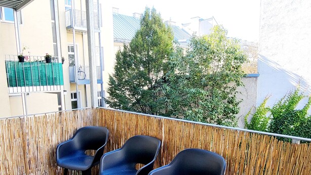 A balcony with three black chairs, a bamboo fence, and a view of buildings and trees. - Siccardsburggasse Apartment With 1 Balcony (Vienna)