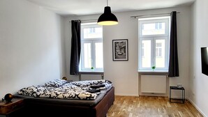 A bedroom with a bed, two windows with curtains, a radiator, a small table with a lamp, and a wall-mounted TV. - Siccardsburggasse Apartment With 1 Balcony (Vienna)